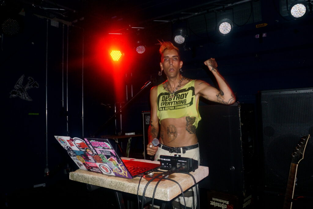 ph4nt. auf der Bühne gegen Ende des Auftritts. Iro hält noch, das neongrüne "Destroy Everything for a better Tomorrow" Croptop ist durchnässt, Gesichtsausdruck grade eher müde. Schweiß läuft über den Bauch. Pose Techno-DJ - Mikro in der Rechten, die Linke zur Faust und halbhochgestreckt